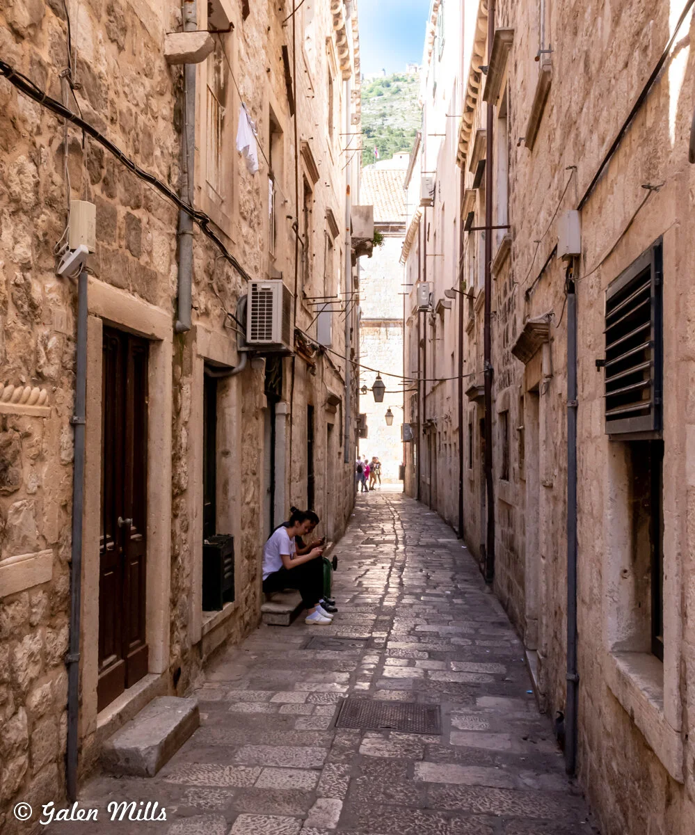 Narrow cobblestone alleyway with stone buildings, person sitting on step using phone, and distant people walking.