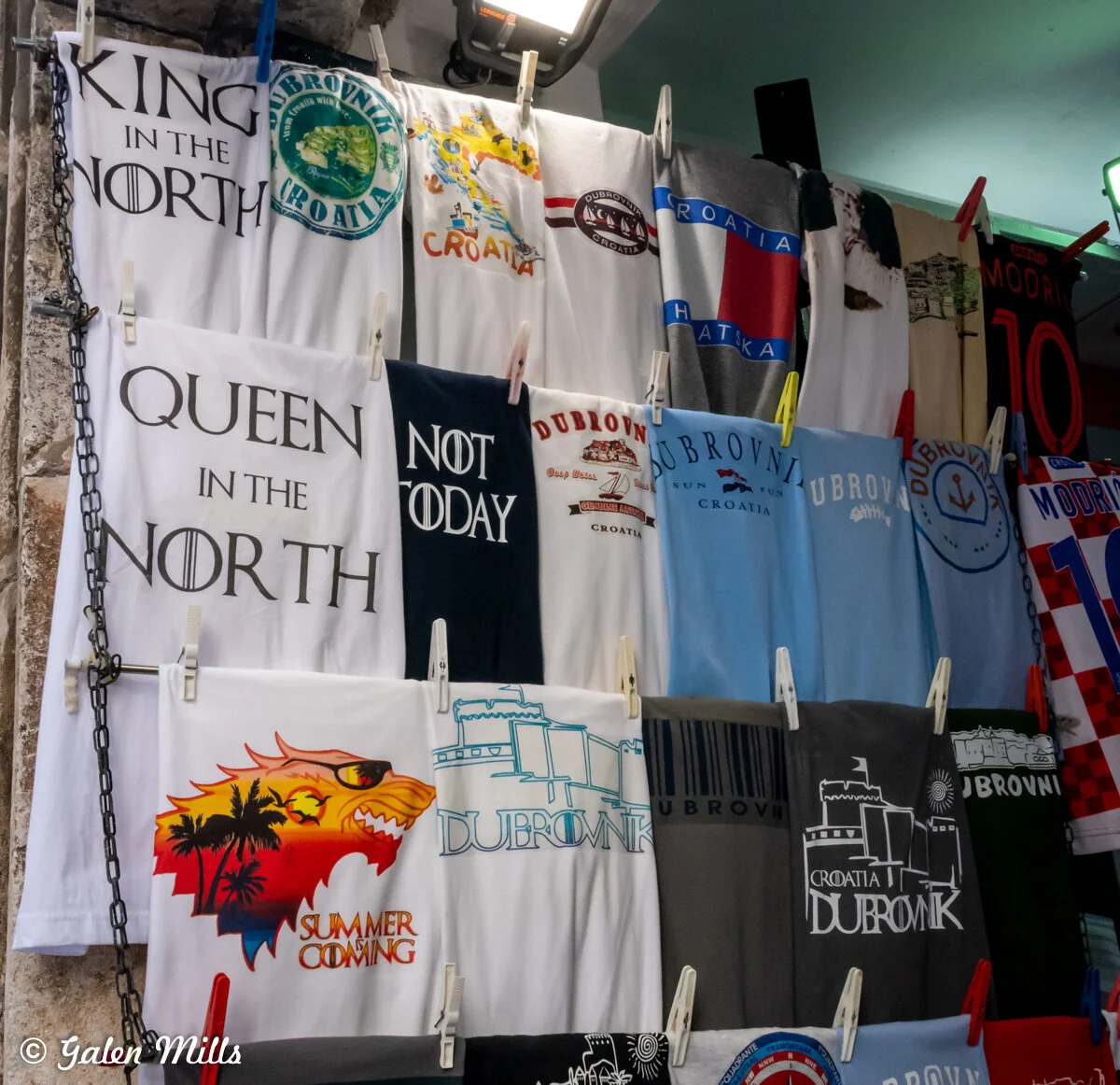 Various Dubrovnik-themed T-shirts displayed on a wall, featuring designs with text such as "King in the North," "Queen in the North," "Not Today," and "Summer Coming," along with Dubrovnik graphics.