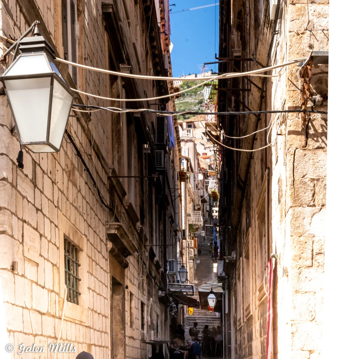 Narrow alleyway with stone buildings and steps in Dubrovnik, Croatia, featuring hanging lamps and visible wires.