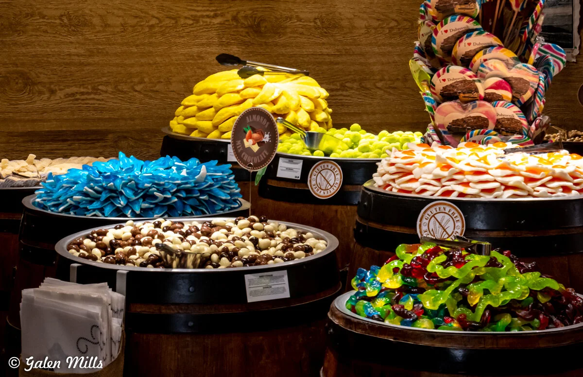 Assorted candies in barrels, including blue shark gummies, chocolate-coated almonds, yellow gummies, and colorful jelly candies, displayed in a confectionery shop.
