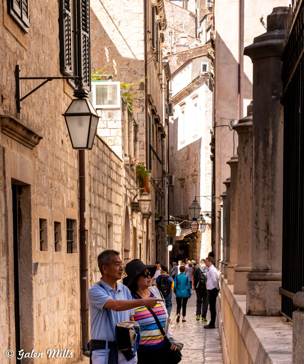 Narrow stone street with tourists in a historic European town, featuring a couple in the foreground, old buildings, street lamps, and other pedestrians.