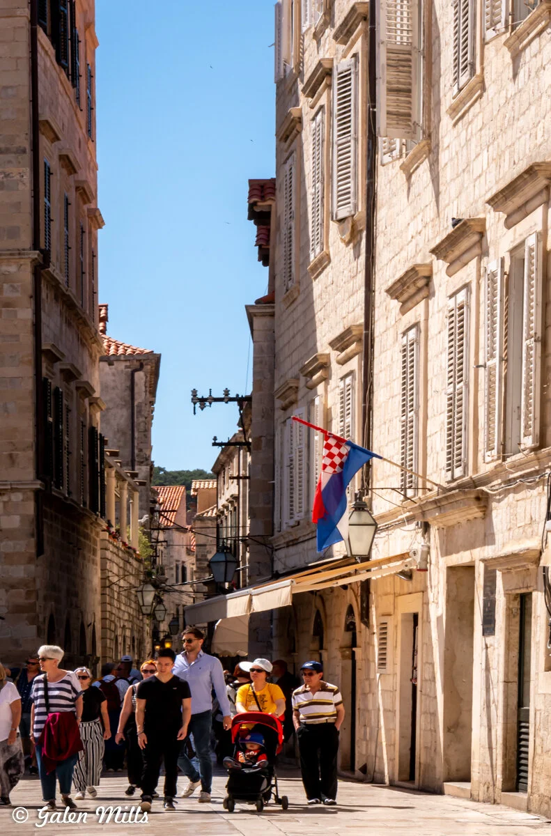 People walking on a narrow stone street in an old European city with historic buildings, featuring a Croatian flag and street lamps.