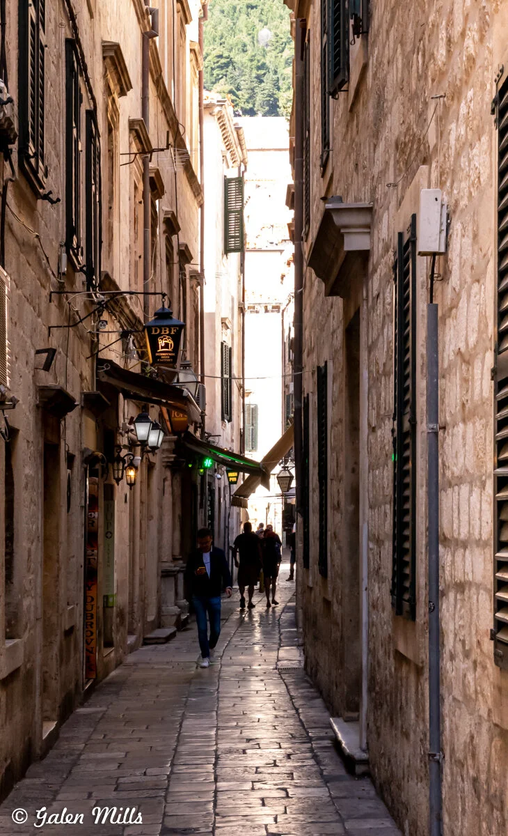 Narrow cobblestone alley with historic stone buildings, vintage signs, and people walking in Dubrovnik, Croatia.
