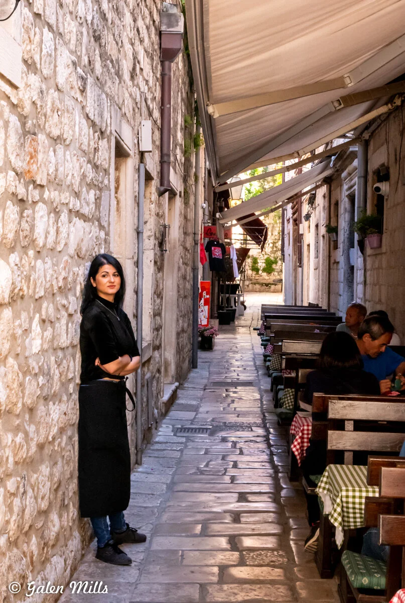 Narrow alley with a person standing by a stone wall, outdoor seating with diners, and tables with checkered tablecloths in a historic setting.