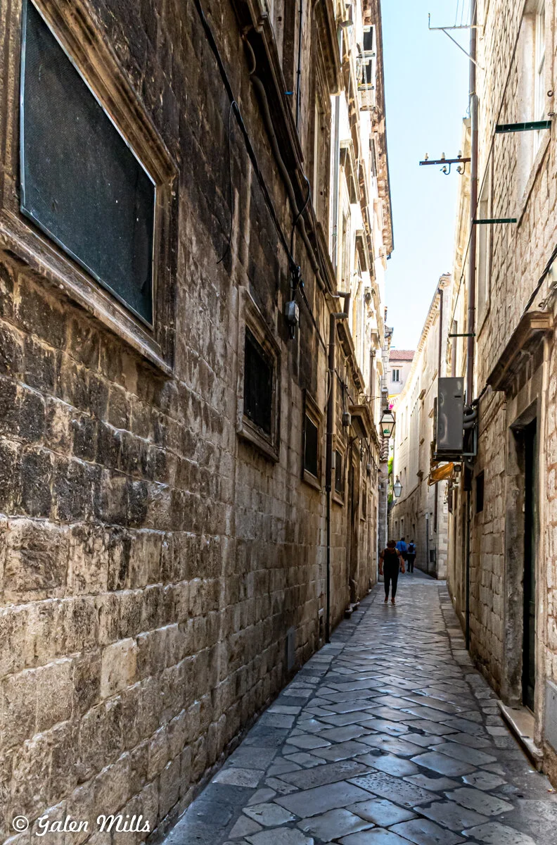 Narrow cobblestone alley with ancient stone buildings on both sides, two people walking, and some balconies.