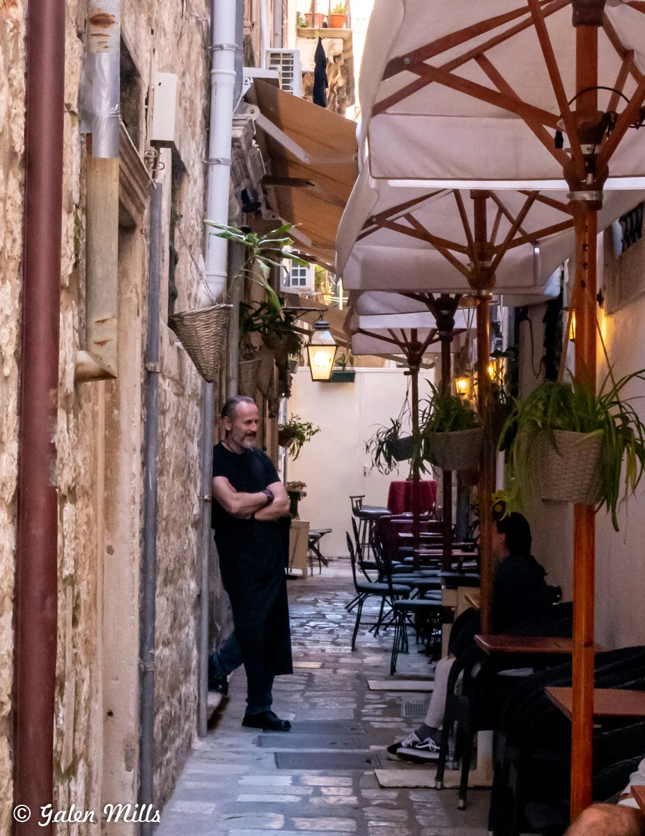 Narrow cobblestone alley with tables and chairs under umbrellas, a man standing with arms crossed, potted plants hanging.