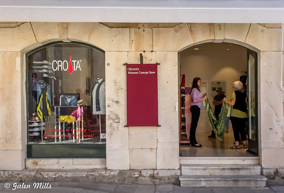 Exterior of Croata Museum Concept Store, featuring a display window with ties and a mannequin, and an open entrance with two women inside engaging with colored fabrics.