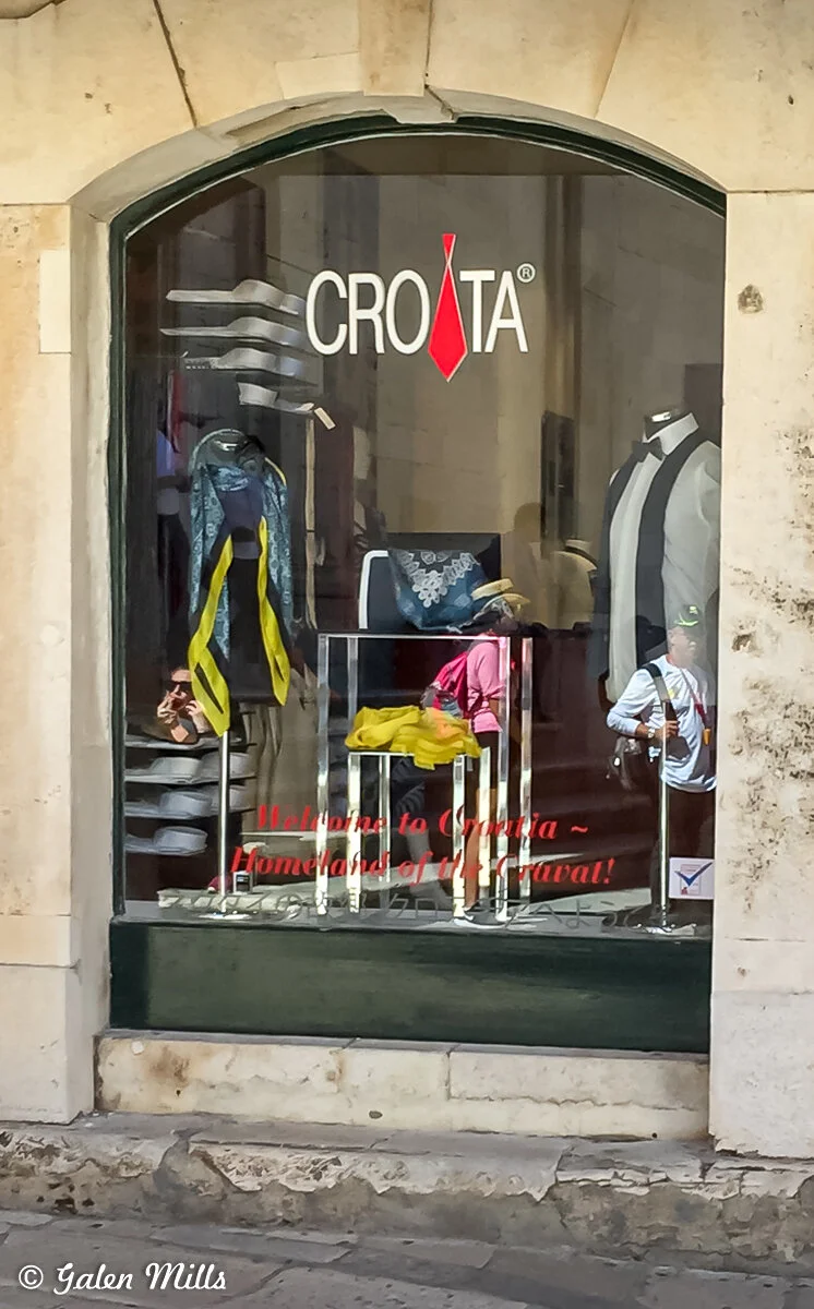 Store window display featuring mannequins dressed in formal attire with ties, various ties on shelves, and a sign reading 'Welcome to Croatia ~ Homeland of the Cravat!' reflecting pedestrians.