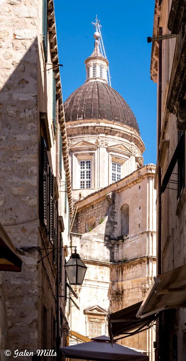 Narrow alleyway with a view of a historic domed building in Dubrovnik, Croatia. The building features a prominent dome, stone walls, and classic architectural details. A street lamp is visible in the foreground.
