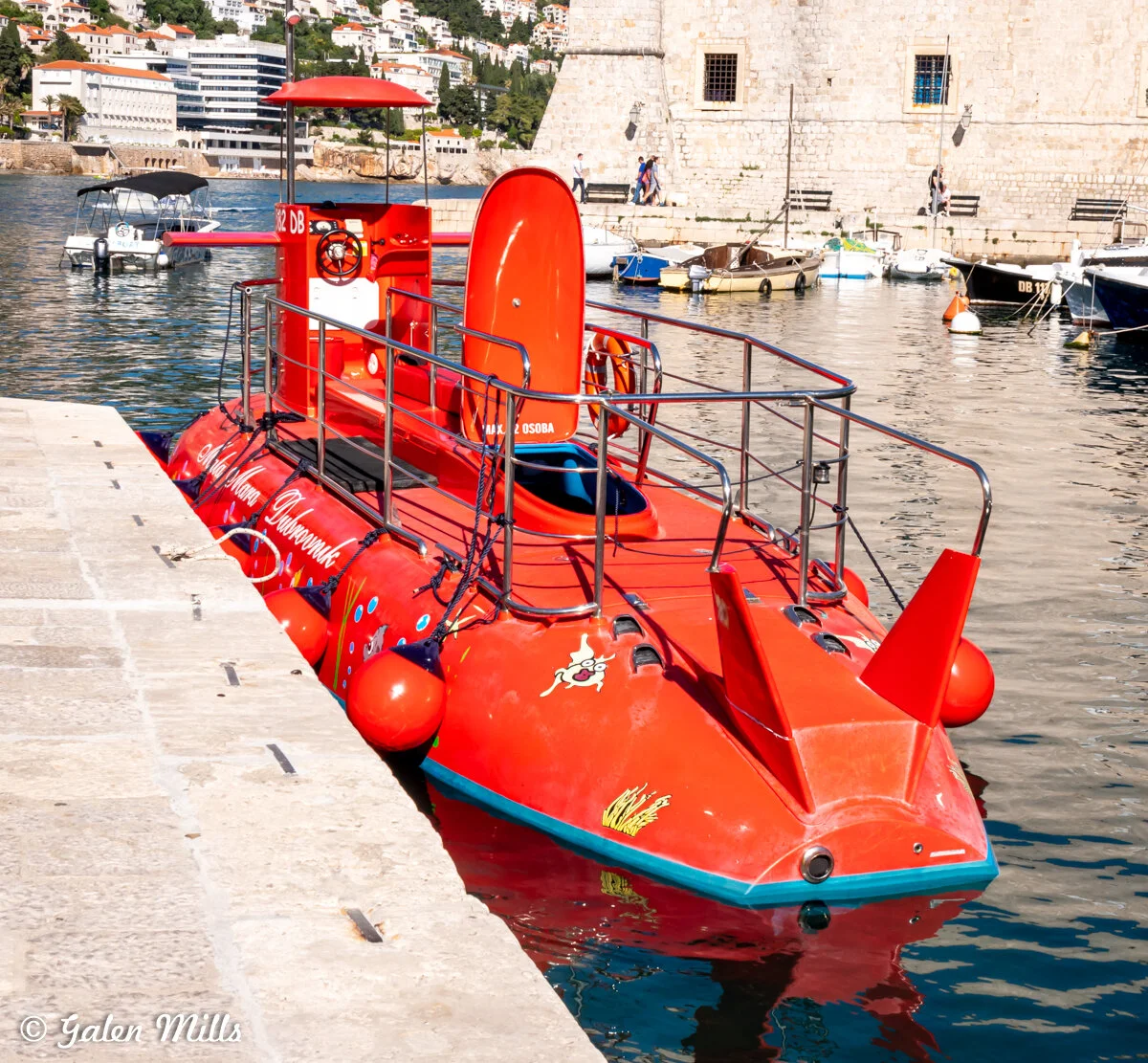 Red submarine tour boat docked at a harbor, featuring safety railings and a closed hatch. The background shows stone buildings, water, and other boats.