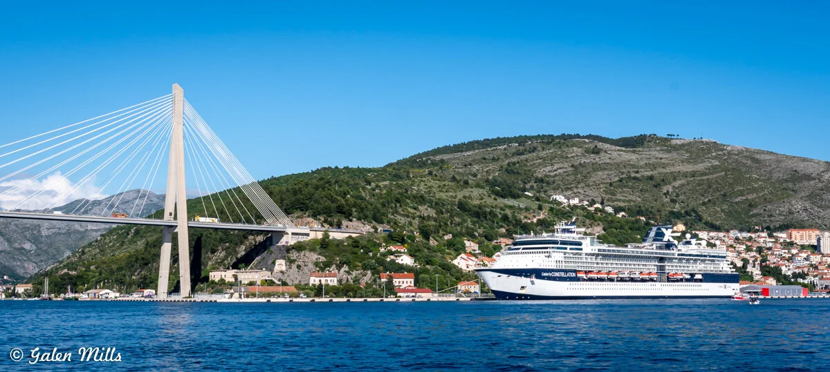 Cruise ship docked near a coastal town, with a cable-stayed bridge and green hills in the background.
