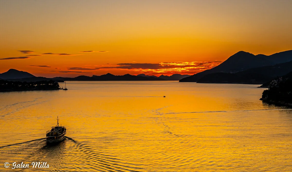 Boat sailing on calm water during a vibrant orange sunset with silhouetted mountains and clouds in the background.