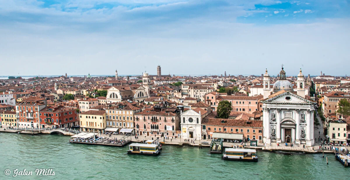 A scenic view of the city of Venice, Italy, showing historic buildings with red roofs and a prominent church with a dome, situated along a canal.