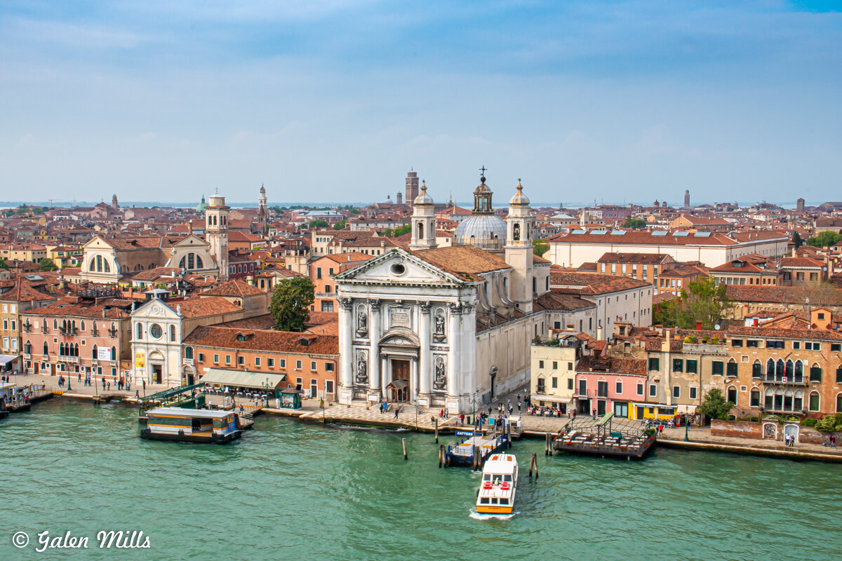 Aerial view of Giudecca Island in Venice featuring a Baroque-style church, surrounding historic buildings with terracotta roofs, in front of a canal with a boat.