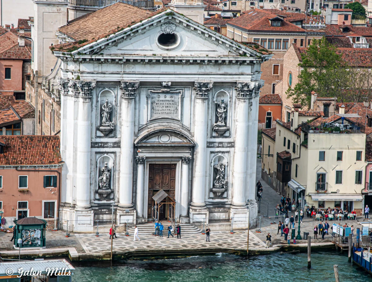 Historical white stone church with statues and columns in Venice, Italy, surrounded by people and water.