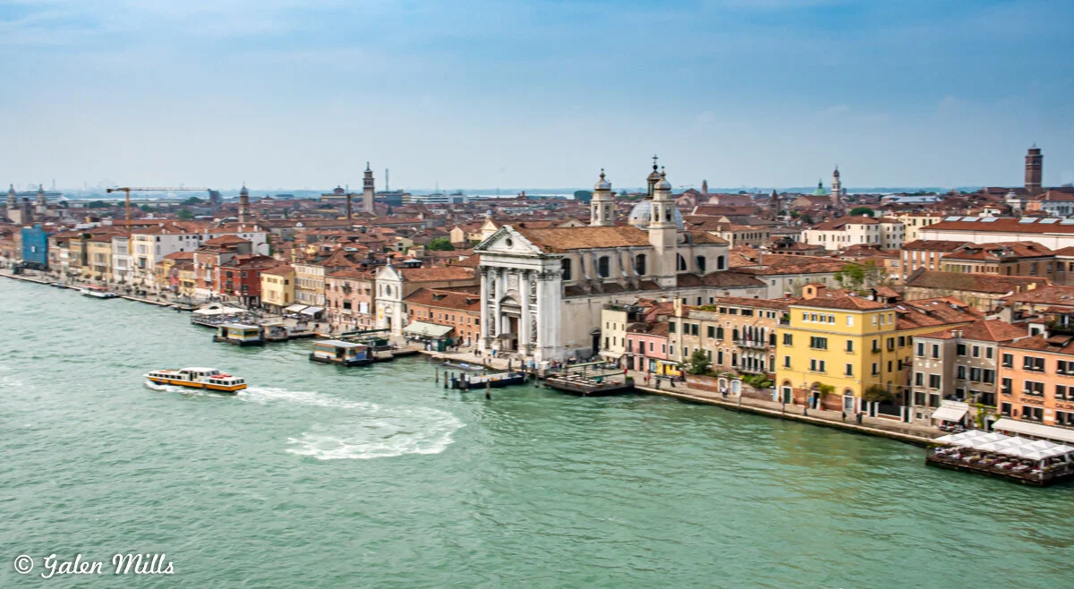 Aerial view of Venice, Italy, featuring a canal lined with colorful buildings, a large historic cathedral, and a passing water taxi.