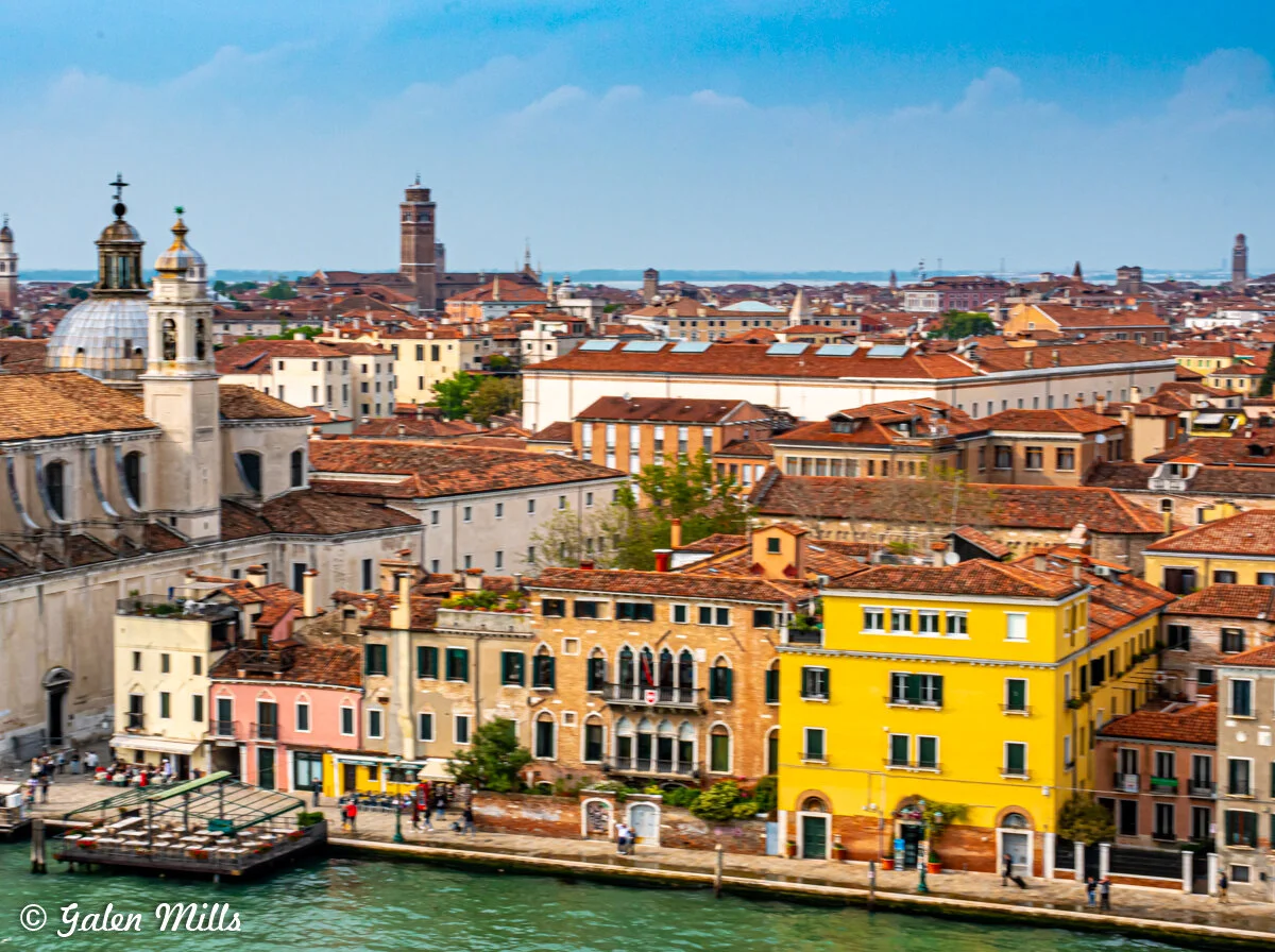 A scenic view of Venice, Italy, featuring colorful buildings along a canal. Prominent architecture with red tile roofs is visible, alongside a domed church and distant towers under a blue sky.
