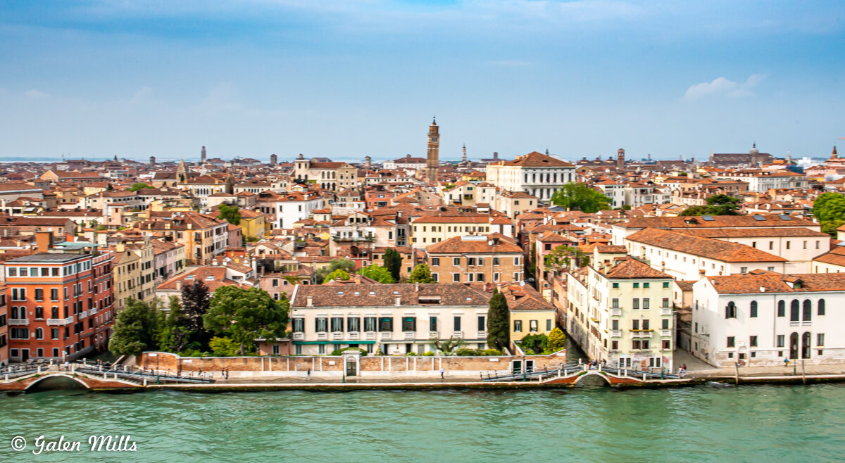 Panoramic view of Venice, Italy with historical buildings, terracotta rooftops, a canal in the foreground, and a clear blue sky.