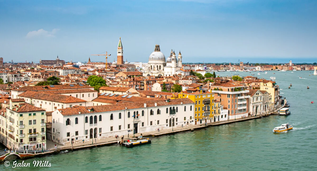 A panoramic view of Venice, Italy, featuring historic buildings with terracotta roofs, a prominent dome, a bell tower, and a canal with boats.
