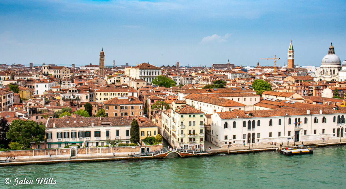 Aerial view of Venice, Italy, featuring historic buildings with terracotta roofs, a canal in the foreground, and notable landmarks such as bell towers and domes under a clear sky.