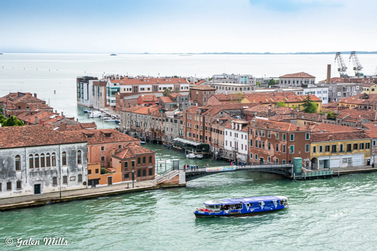 Aerial view of Venice with a canal and colorful buildings, featuring a blue boat in the foreground, against a backdrop of the sea and a clear sky.
