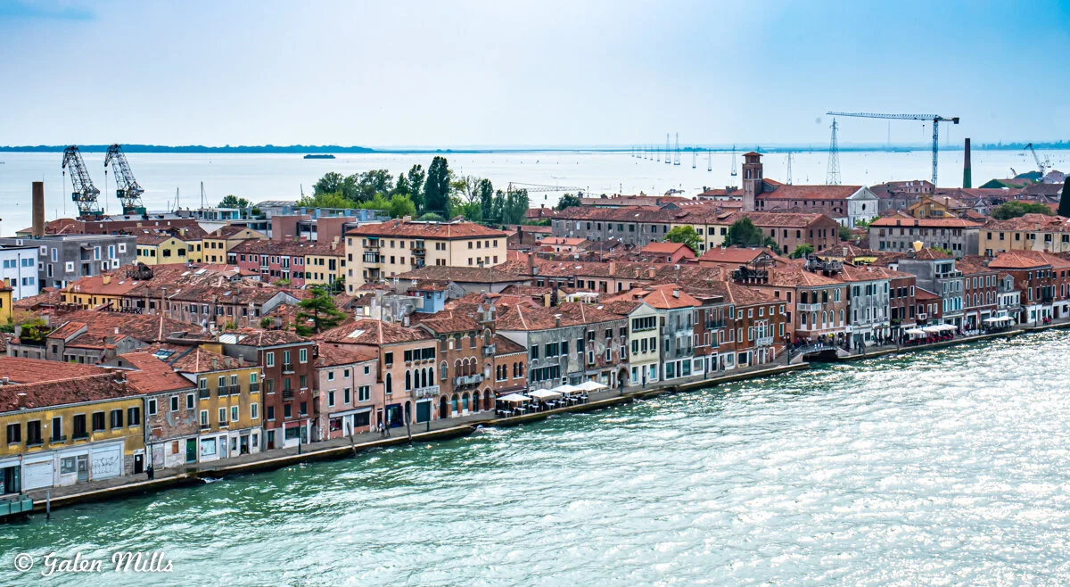 Aerial view of colorful buildings lining a canal in Venice, Italy, with cranes and the distant shoreline in the background.