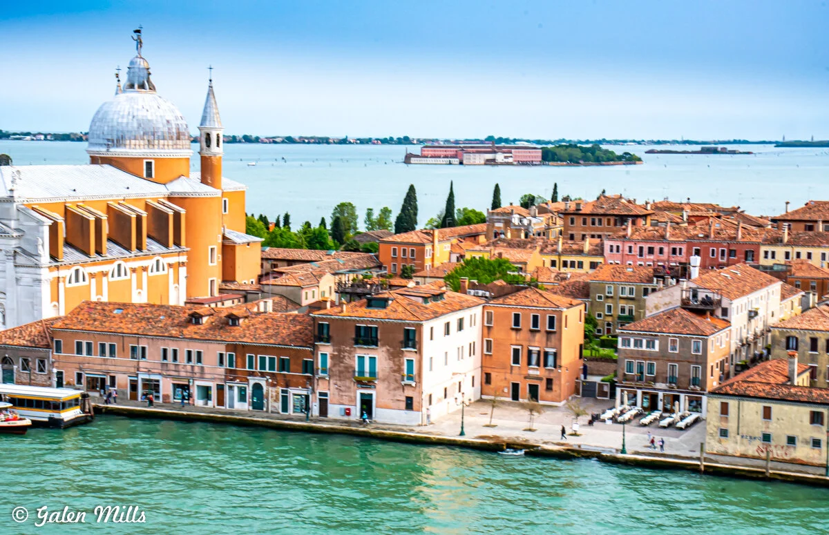 Aerial view of Venice, Italy, featuring a large domed church near the waterfront, surrounded by colorful historic buildings and a canal. A distant island and blue sky are in the background.