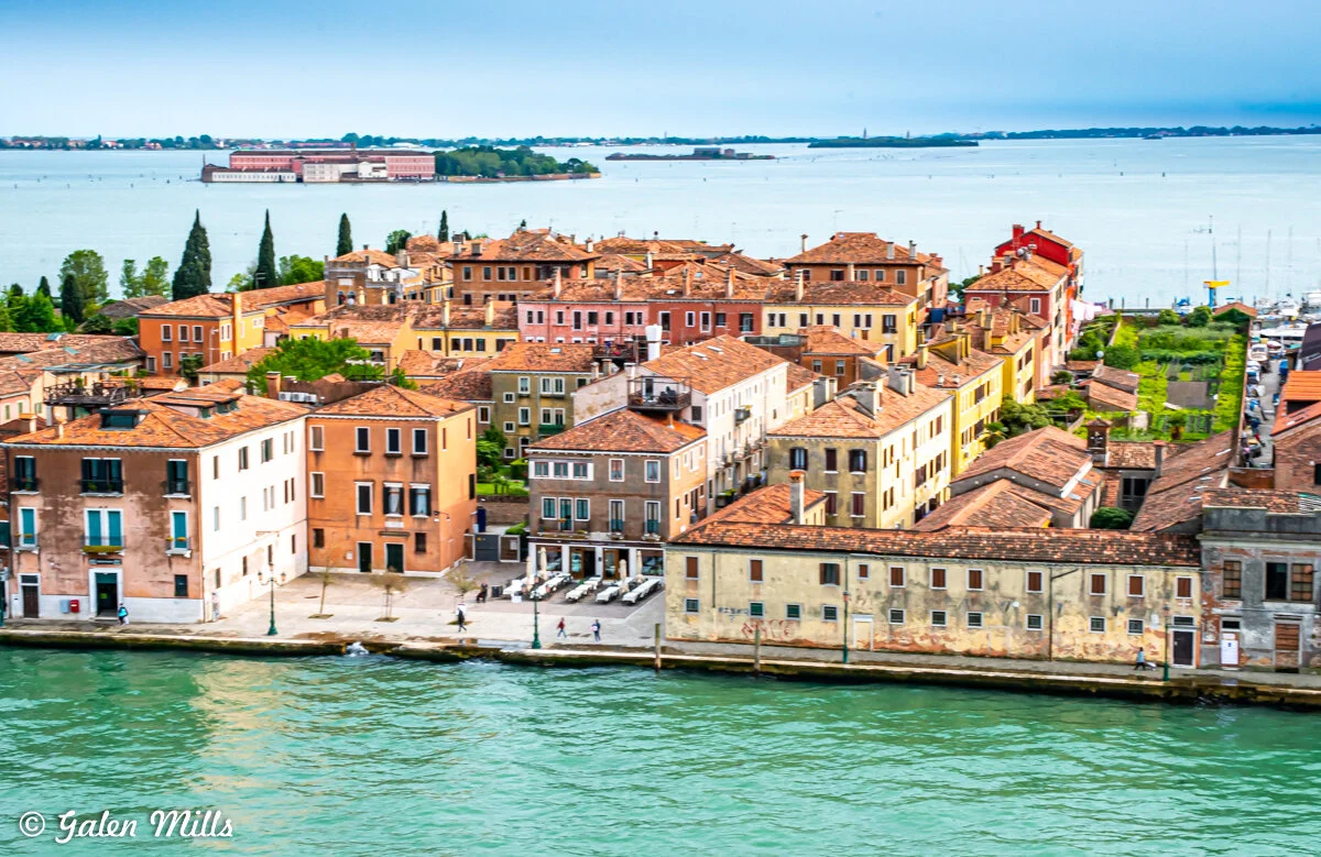 Aerial view of colorful, historical buildings with terracotta roofs along a waterfront in Venice, Italy, set against a backdrop of green trees and a blue lagoon under a cloudy sky.