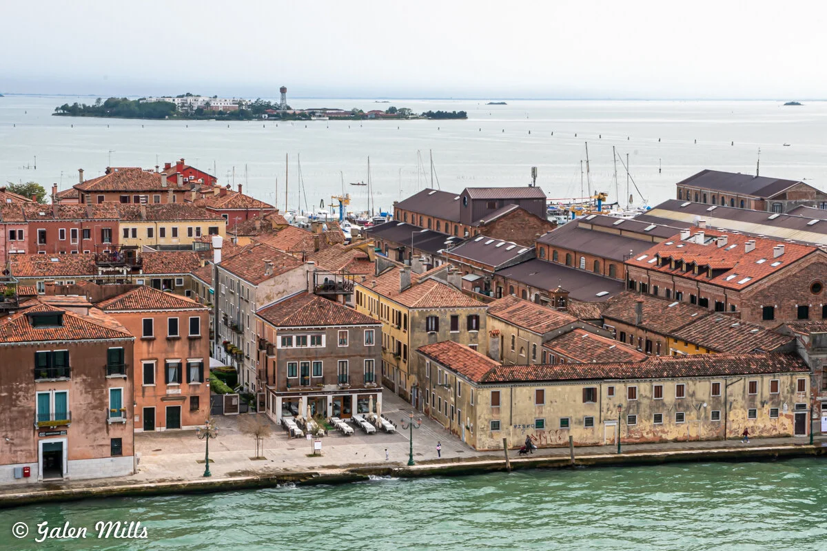 Aerial view of Venice waterfront with colorful historic buildings, red-tiled roofs, and a canal. Boats are moored in the background, with an island visible across the water.