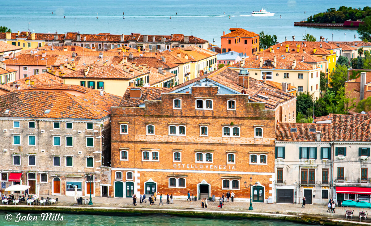 View of Venice with brick buildings, a canal, and people walking along the waterfront. In the background, a boat is sailing on the water.