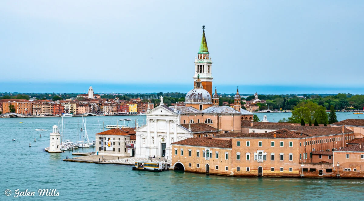 Aerial view of San Giorgio Maggiore island in Venice, Italy, featuring a large church with a prominent bell tower and dome, surrounded by water with the cityscape in the background.