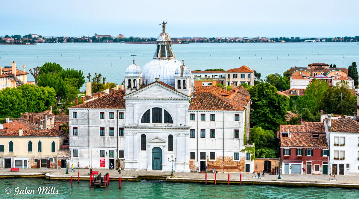 Historic church building in Venice, Italy, with a large dome, set along a waterfront with trees and colorful rooftops. In the background, a wide expanse of water is visible.