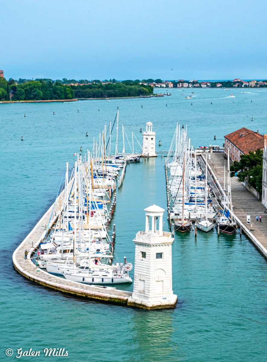 Scenic view of a marina with sailboats docked along a harbor, flanked by a white lighthouse. The background shows a calm turquoise sea, a distant shoreline with trees, and scattered buildings under a blue sky.