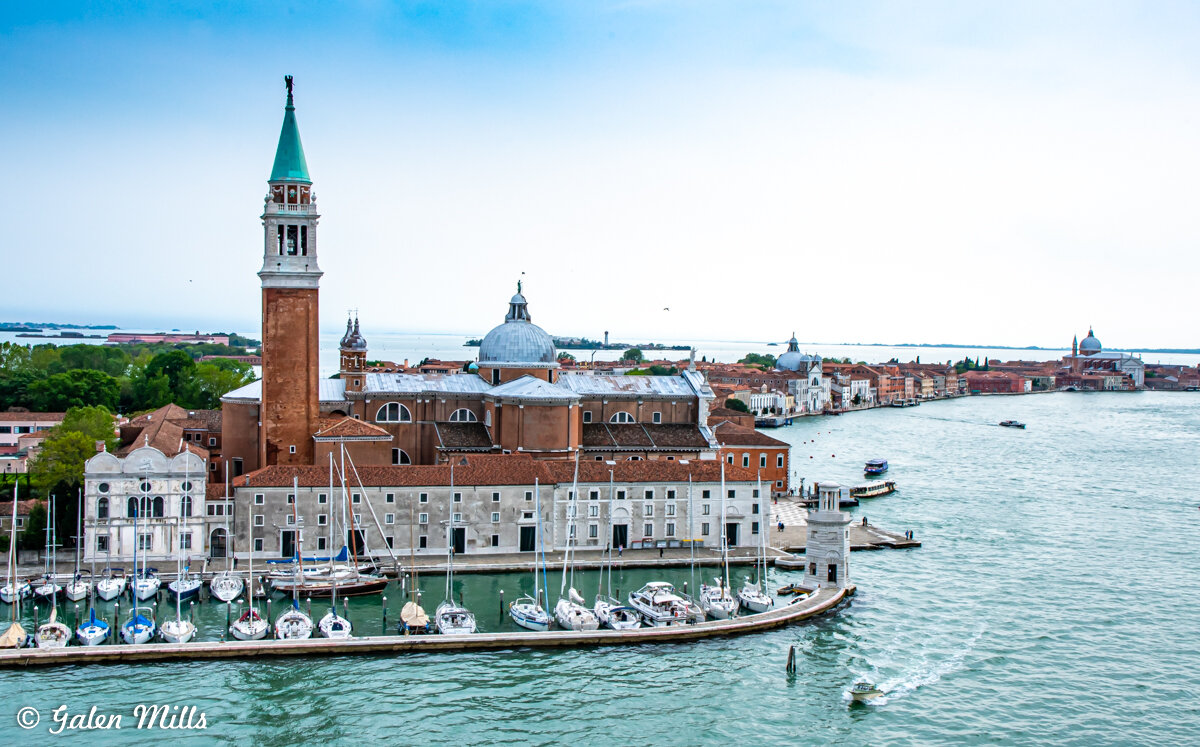 View of San Giorgio Maggiore island in Venice, featuring a prominent church with a tall bell tower, boats docked along the marina, and the surrounding lagoon.