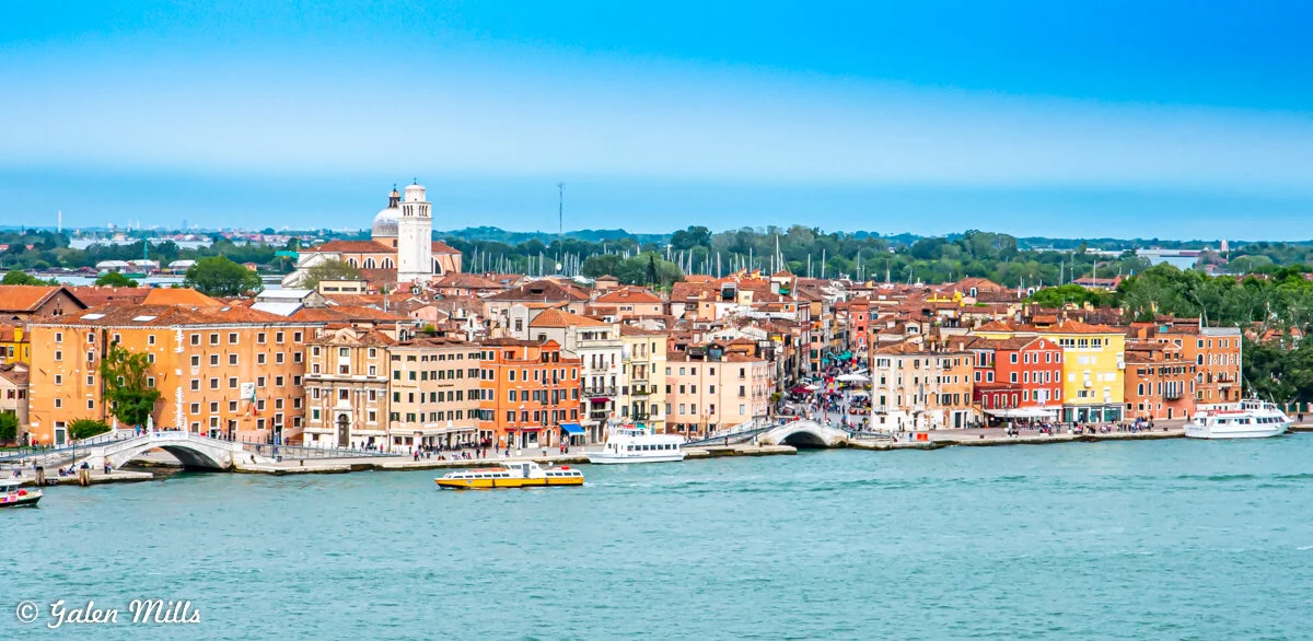 Scenic view of a colorful waterfront city with bridges, boats, and a church dome, surrounded by green trees and a clear blue sky.