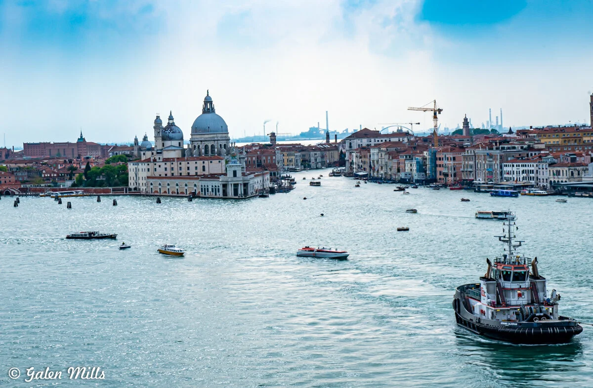 View of Venice's Grand Canal with historic buildings, including a domed church, boats, and a distant industrial skyline.