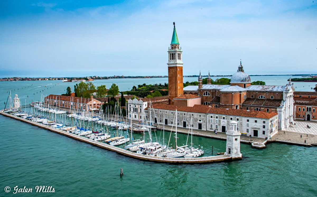 Aerial view of San Giorgio Maggiore island in Venice, Italy, featuring the church with its tall bell tower and a marina full of sailboats on the turquoise water.