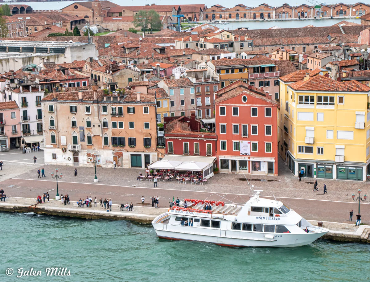 Scenic view of a canal in Venice with docked white ferry, colorful buildings, café with outdoor seating, people walking along the promenade, and historic architecture in the background.