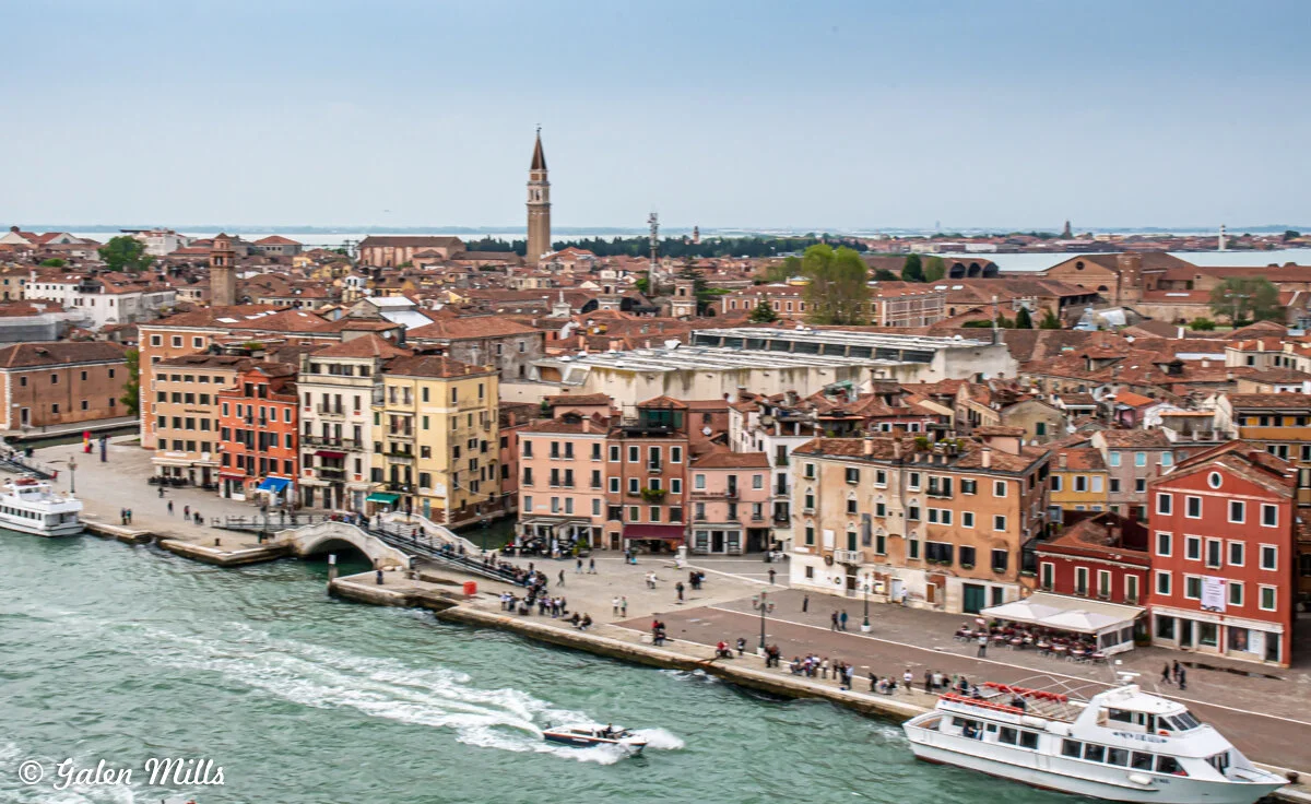 Aerial view of historic buildings and boats in Venice, Italy, near a waterfront with people walking along the promenade.