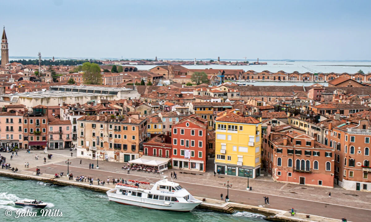 Aerial view of colorful buildings along the waterfront in Venice, Italy, with a white boat in the canal and people walking on the promenade.