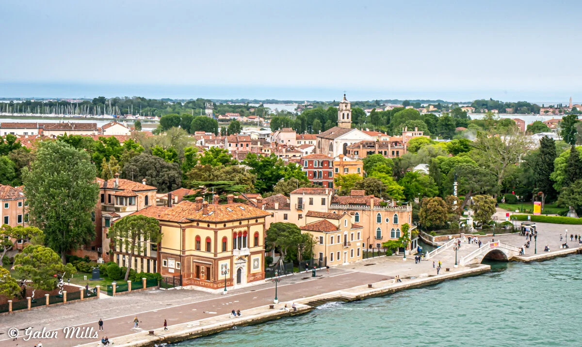 Aerial view of Venice, Italy with buildings, trees, and a waterfront walkway. A church with a bell tower is in the background, and there is a bridge over a canal. The area is surrounded by greenery and adjacent waterways.