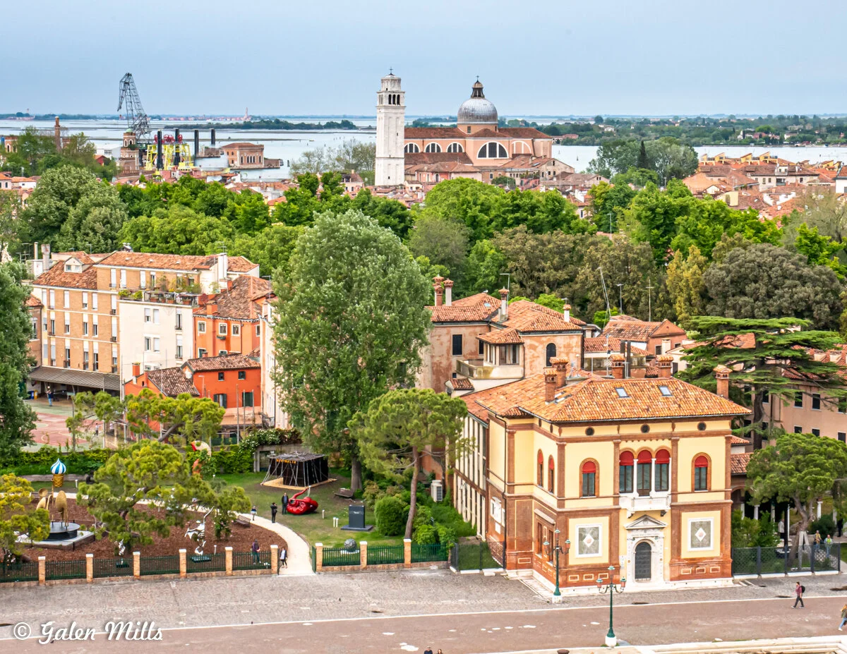 Aerial view of Venice showing traditional buildings with terracotta roofs, lush greenery, and a domed church. The cityscape is bordered by a body of water in the background, along with industrial cranes on the horizon.