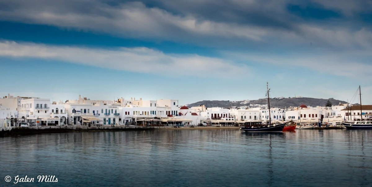 Harbor view of white buildings with blue doors, small boats moored, and calm water in Mykonos, Greece.