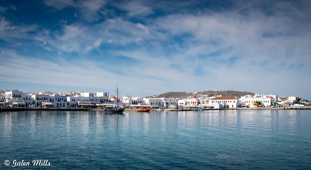 Waterfront view of a coastal town with white buildings and boats in the harbor under a blue sky.