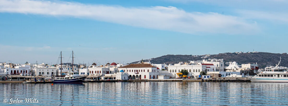 Waterfront view of a coastal town with white buildings, boats docked at the pier, and hills in the background.