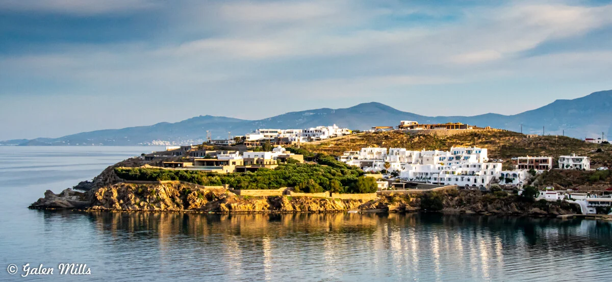 Scenic view of white buildings on an oceanfront hillside, surrounded by greenery, with a calm sea and distant mountains under a cloudy sky.