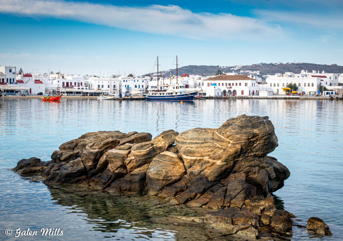 Rocks in clear water with a distant view of a waterfront town featuring white buildings and boats in a harbor under a blue sky.
