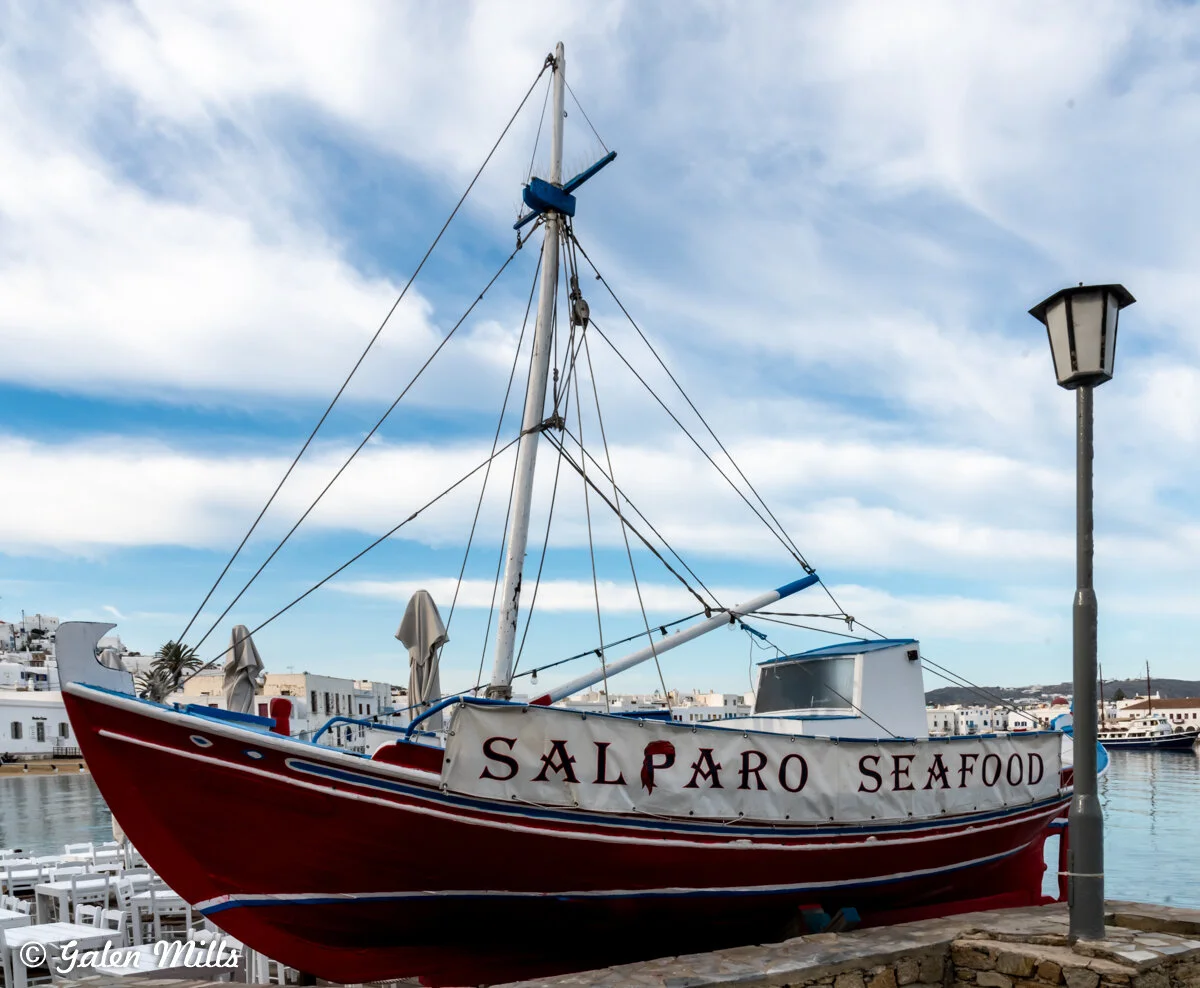 A red and white boat with "Salparo Seafood" written on its side, moored by a waterfront with a lamppost nearby, against a backdrop of a harbor and buildings.