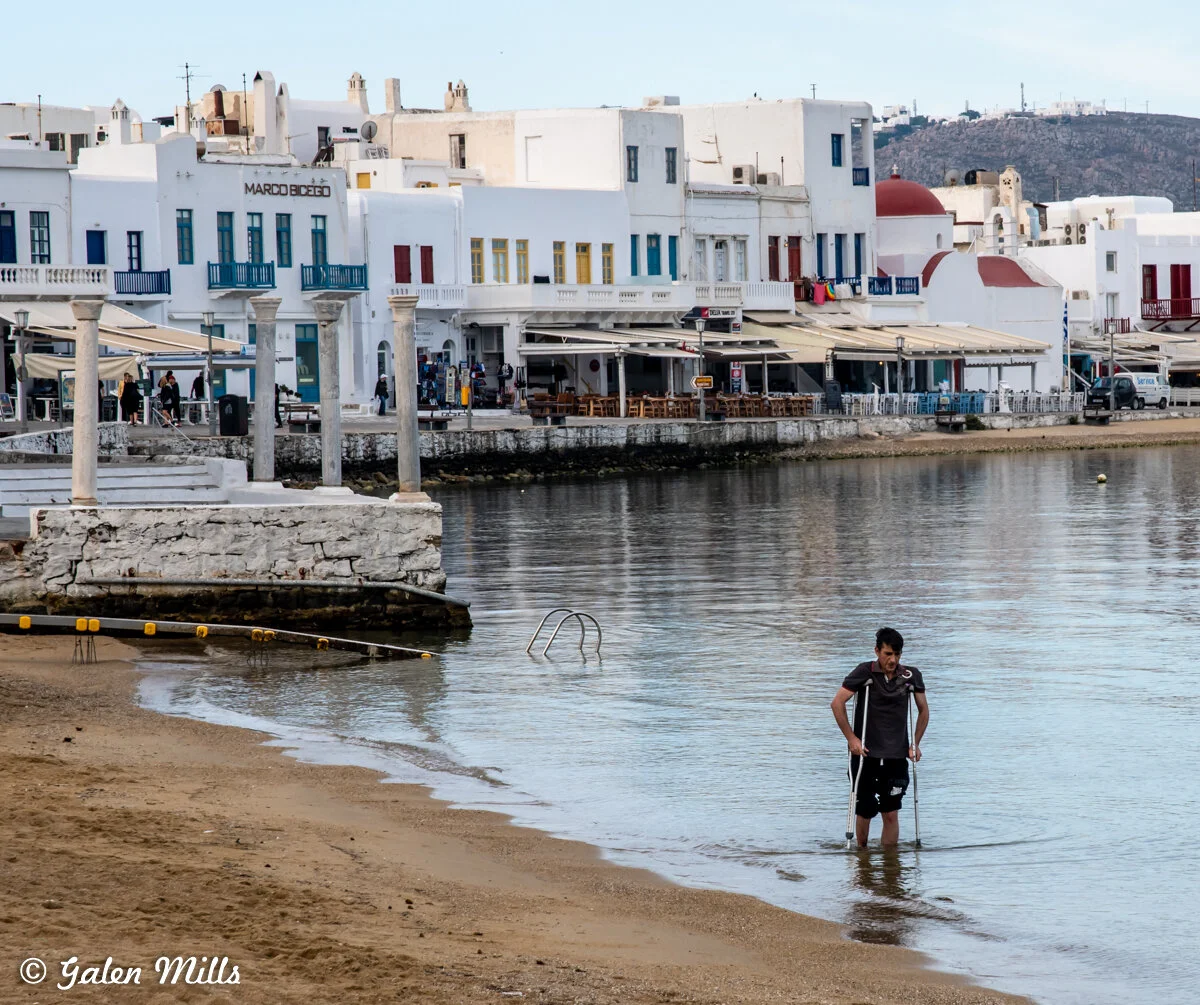 Man with crutches walking in shallow water near a sandy beach, with white buildings and outdoor cafes in the background.