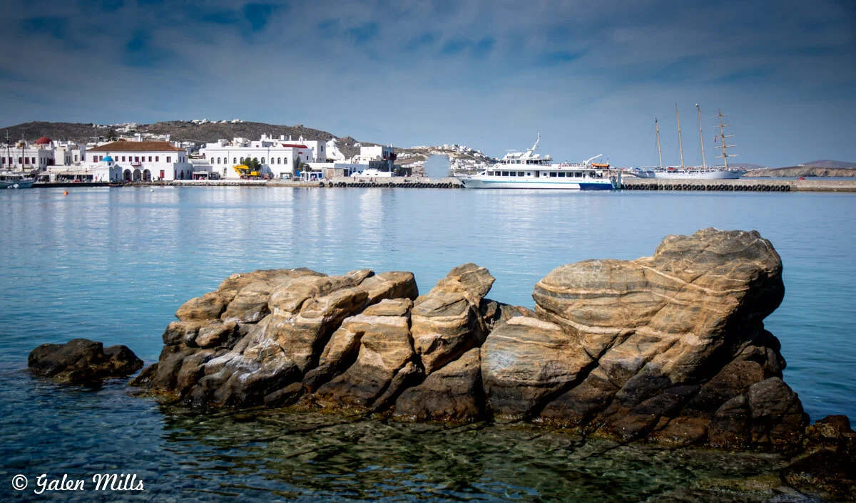 Rocky shoreline with a coastal town in the background, featuring white buildings and boats anchored at a harbor.